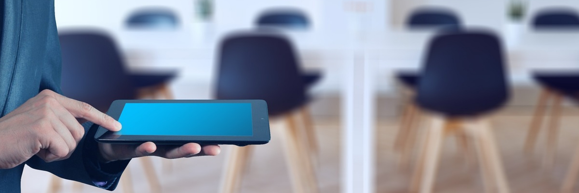 Jemand hat ein Tablet in der Linken Hand und tippt mit dem rechten Zeigefinger daraum. Im Hintergrund sind Tische und Stühle verschwommen zu erkennen.